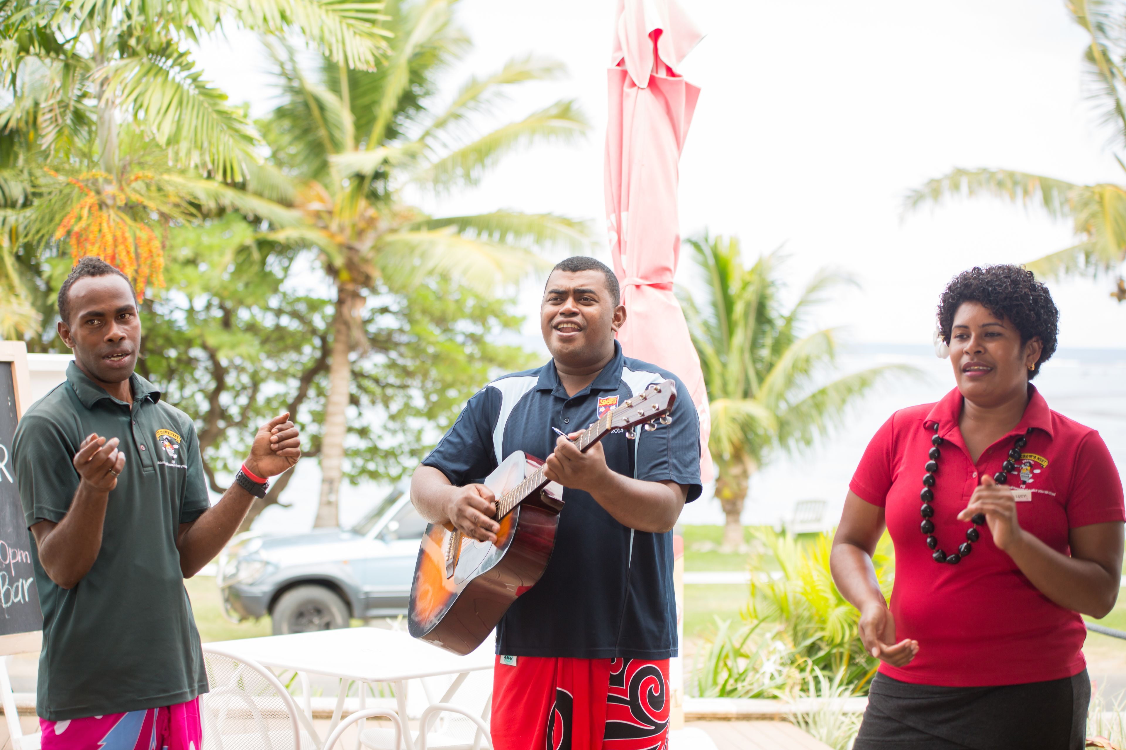 Three people standing outdoors, one playing a guitar, with palm trees and a car in the background.