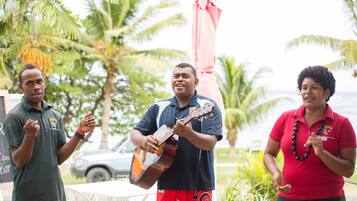 Three people standing outdoors, one playing a guitar, with palm trees and a car in the background.