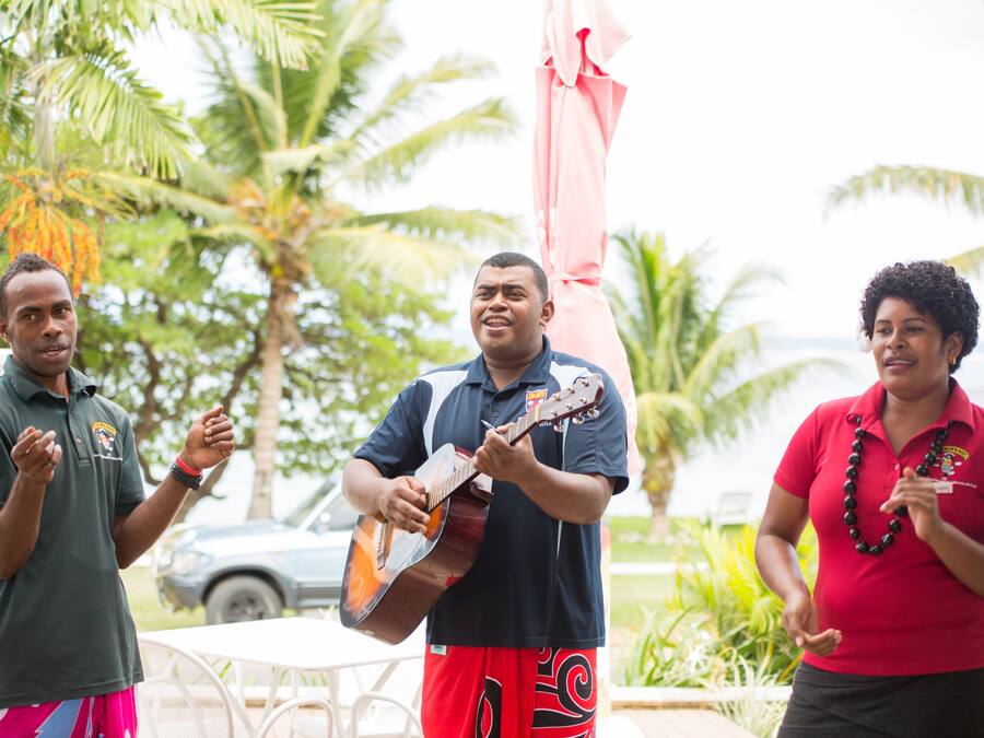 Three people standing outdoors, one playing a guitar, with palm trees and a car in the background.