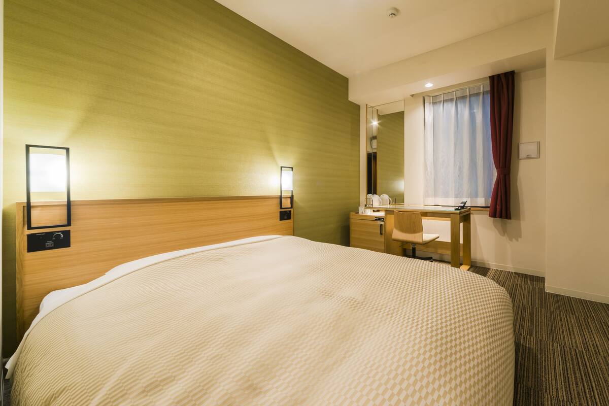 Ein Hotelzimmer mit einem großen Bett, einem Schreibtisch mit Stuhl, einem Spiegel, einem Fernseher und einem Fenster mit Vorhängen.