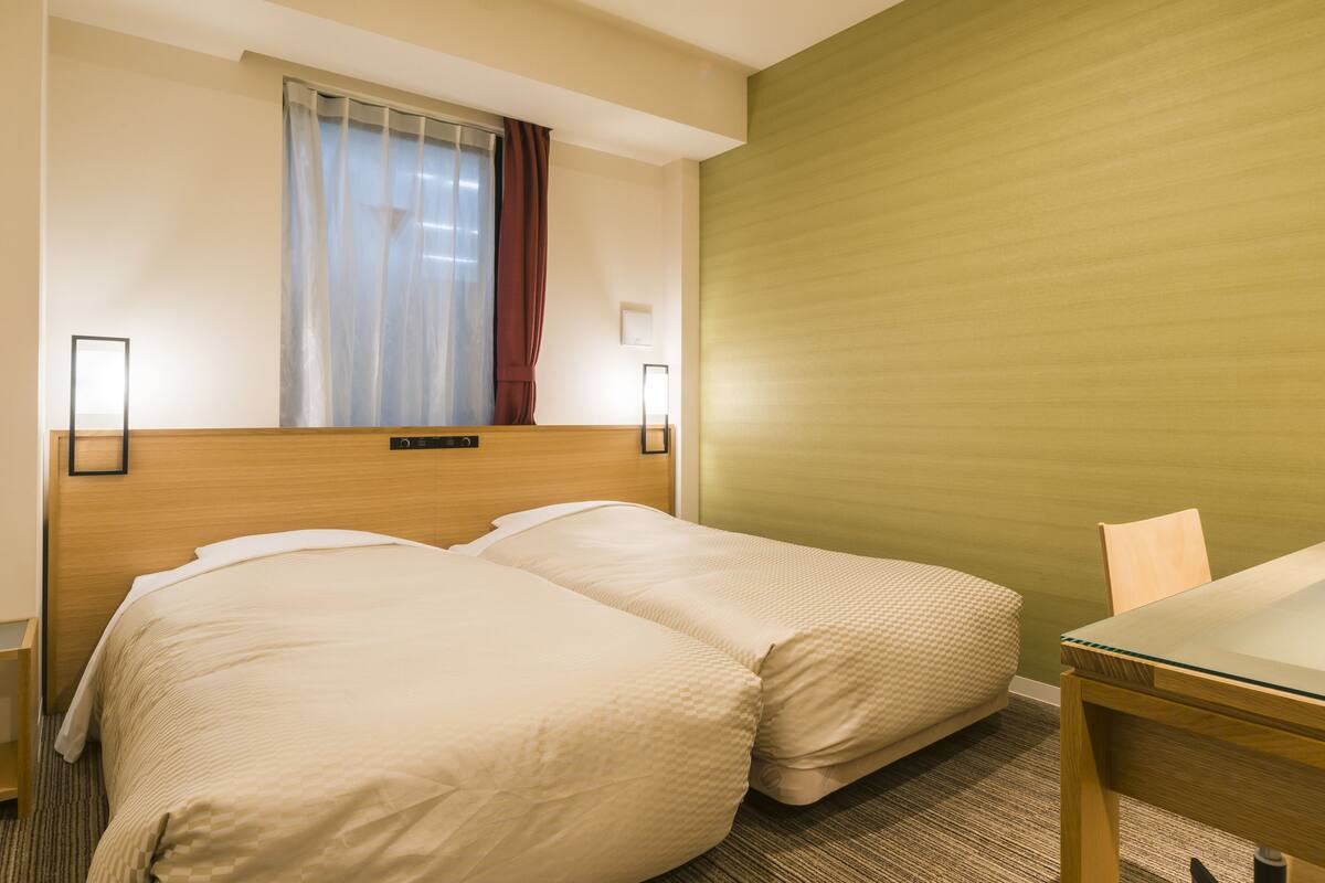 Ein Hotelzimmer mit zwei Betten, einem Holz-Kopfteil, einem Fenster mit Vorhängen, einem Nachttisch und einem Stuhl.