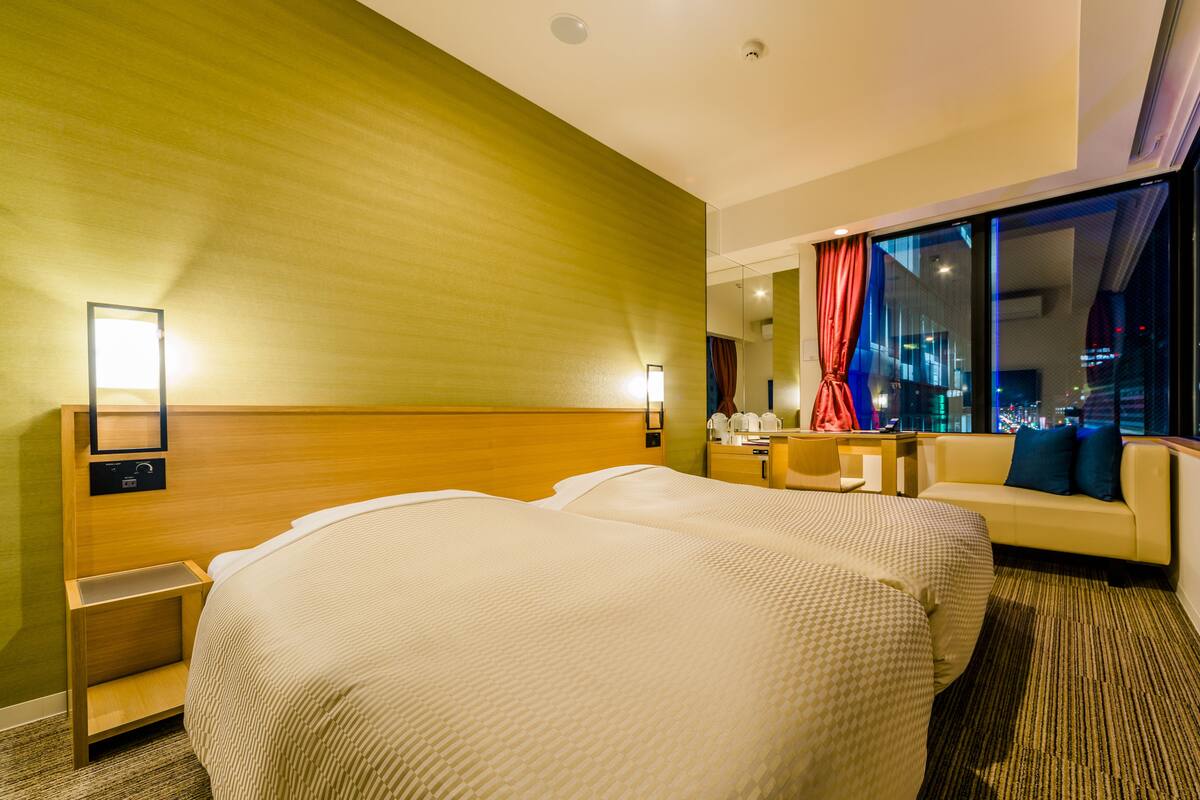 Ein Hotelzimmer mit zwei Betten, einem Schlafsofa, einem Schreibtisch und einem großen Fenster mit Blick auf die Stadt.