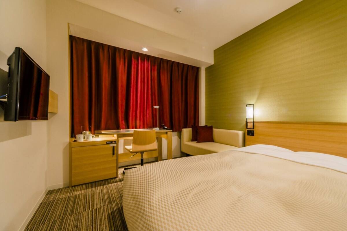 Ein Hotelzimmer mit Bett, Schreibtisch, Sessel, Fernseher und einem großen Fenster mit roten Vorhängen.