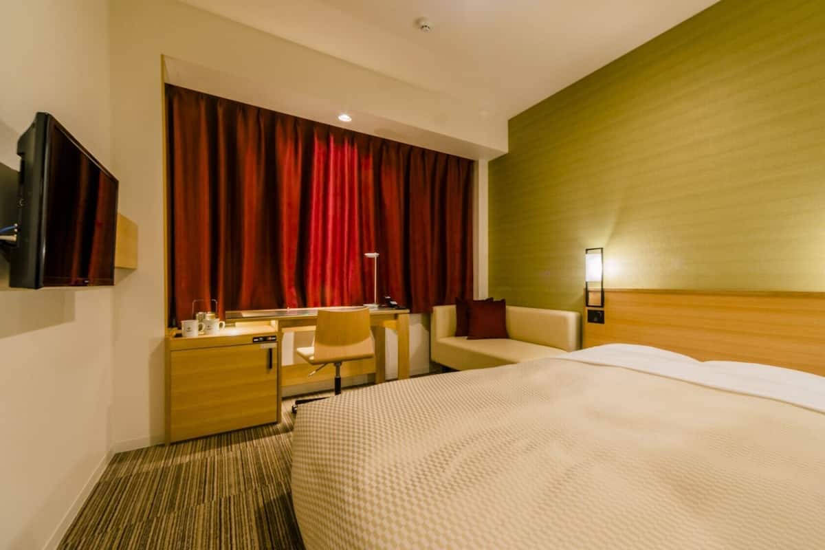 A hotel room with a bed, a desk, a chair, a TV, and a large window with red curtains.