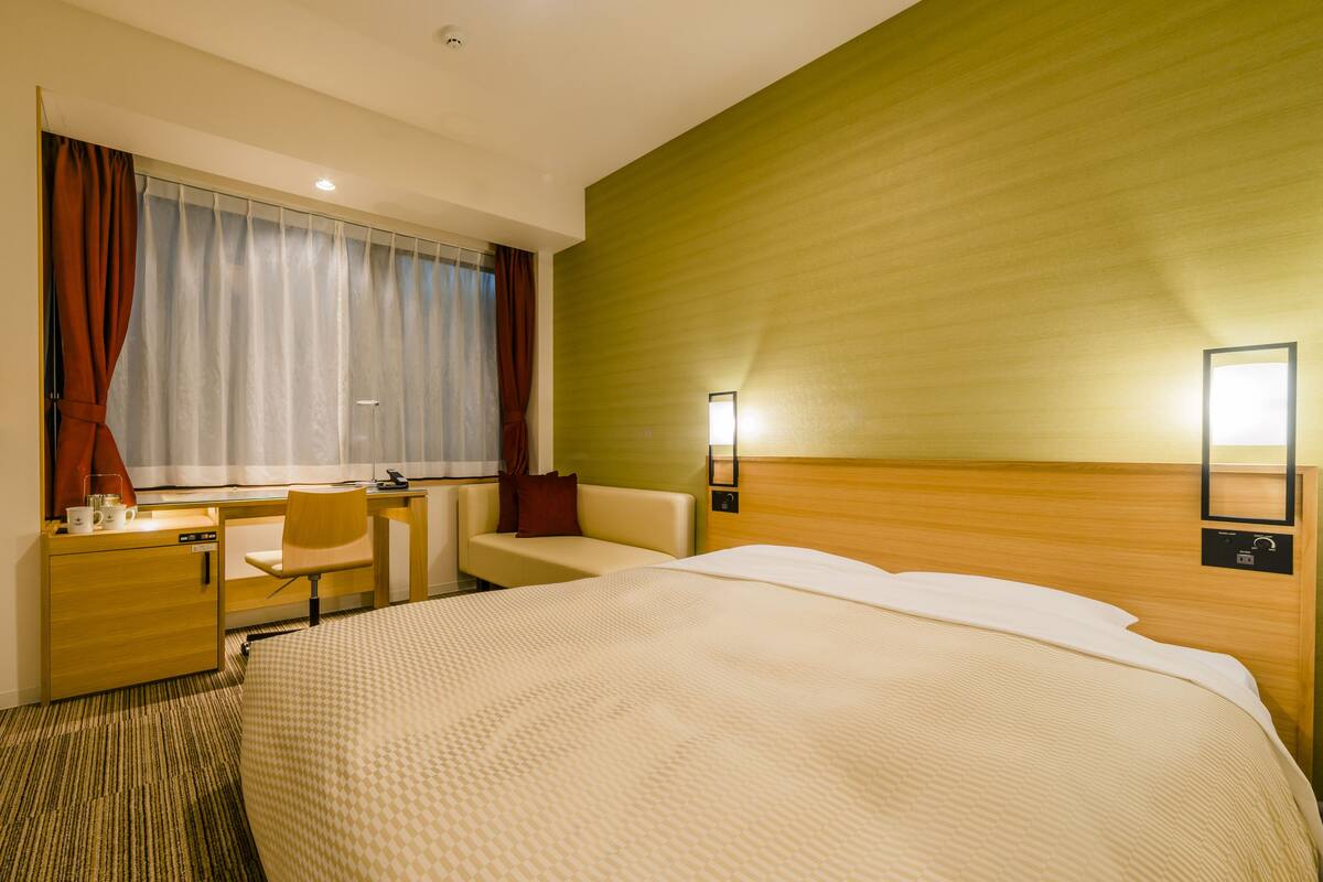 Ein Hotelzimmer mit Bett, Schreibtisch mit Stuhl, Fernseher, Fenster mit Vorhängen und einer Wandleuchte.