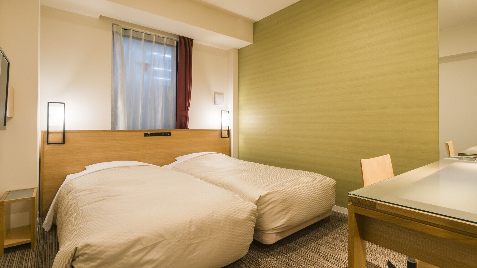 A hotel room with two beds, a wooden headboard, a window with curtains, a green accent wall, a small table, and two wall-mounted lamps.
