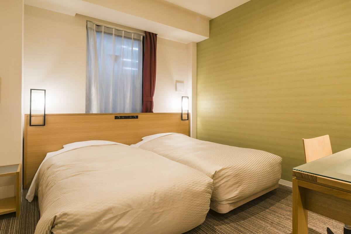 Ein Hotelzimmer mit zwei Betten, einem Holzkopfende, einem Fenster mit Vorhängen, einer grünen Akzentwand, einem kleinen Tisch und zwei Wandleuchten.