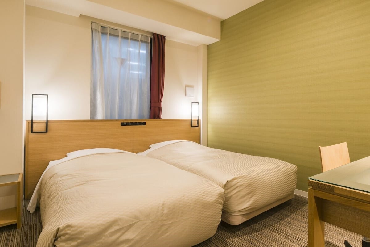 A hotel room with two beds, a wooden headboard, a window with curtains, a green accent wall, a small table, and two wall-mounted lamps.