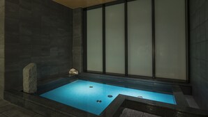 A modern spa room with a rectangular hot tub, dark stone walls, and a large frosted glass window.