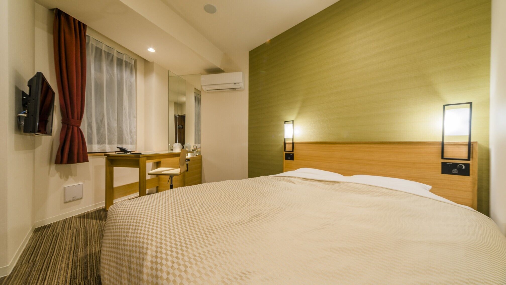 A hotel room with a large bed, a desk, a chair, a mirror, an air conditioning unit, and two wall-mounted lights.