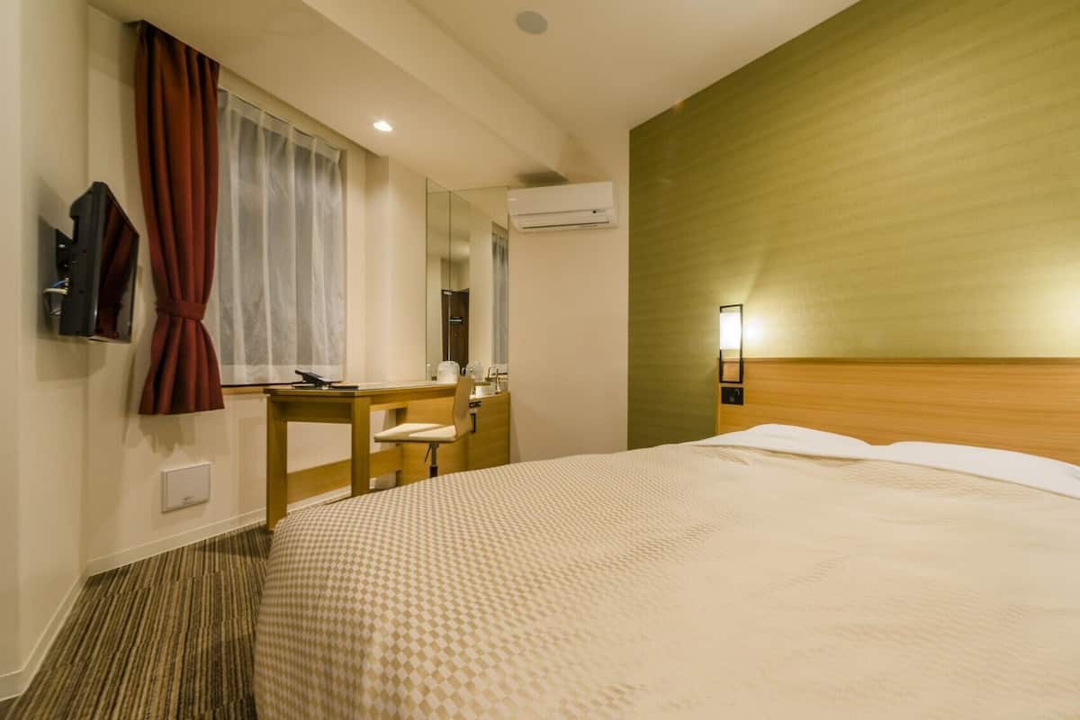 A hotel room with a large bed, a desk, a chair, a mirror, an air conditioning unit, and two wall-mounted lights.