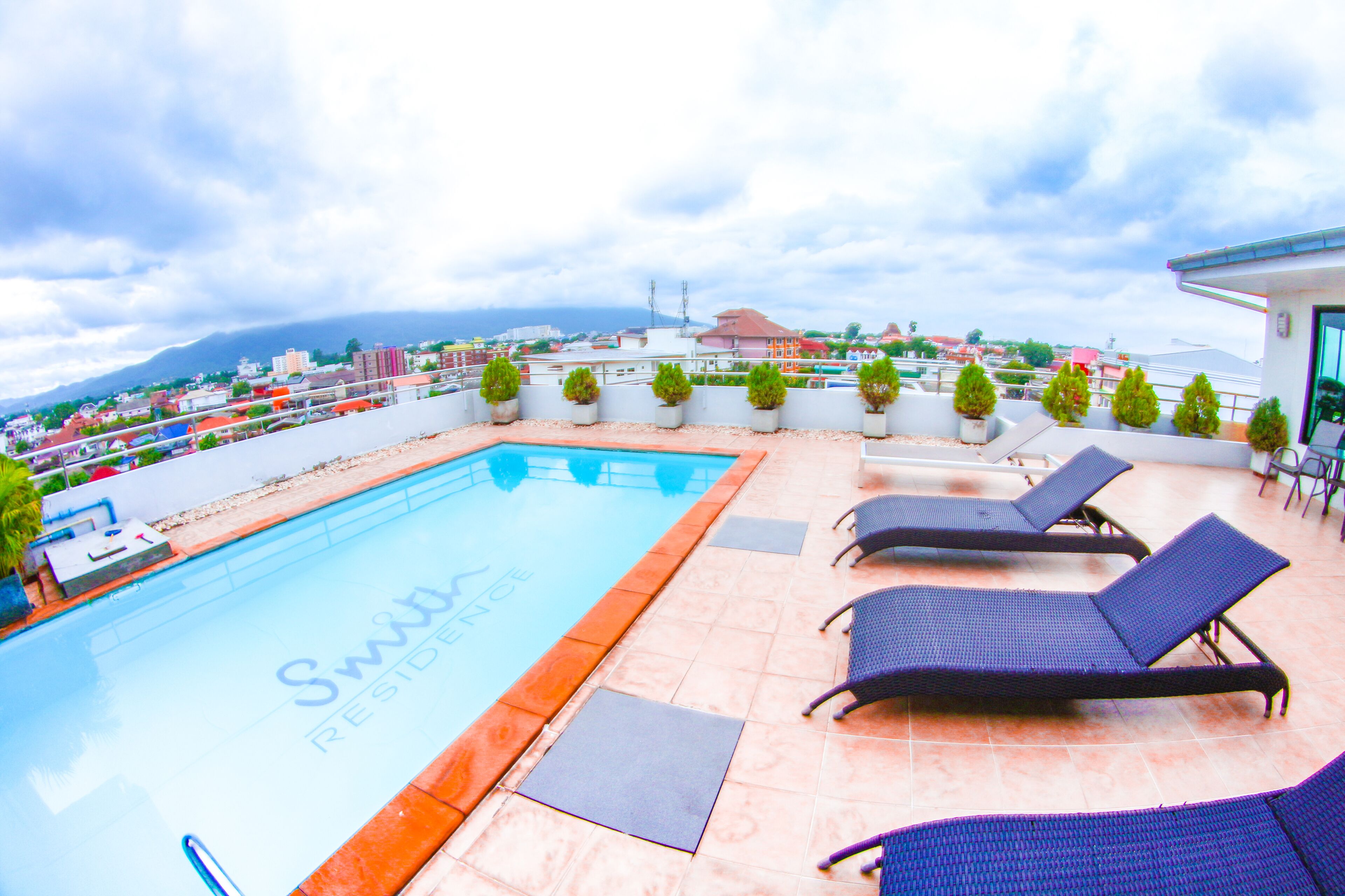 A rooftop pool area with lounge chairs and potted plants, overlooking a cityscape.
