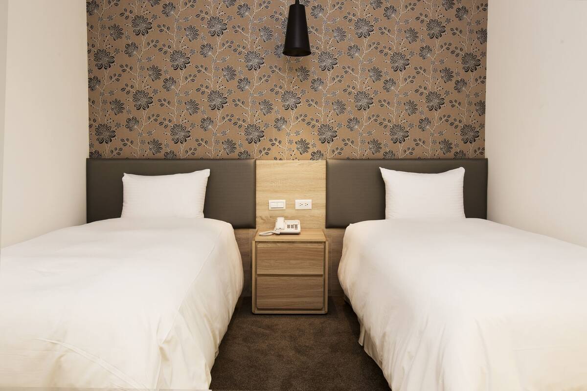 A hotel room with two single beds, a wooden nightstand with a phone, and a floral-patterned headboard.