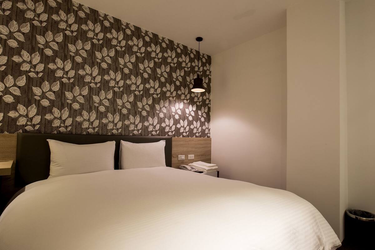 A hotel room with a large bed, a headboard with a leaf pattern, a bedside table with a phone, and a wall-mounted light fixture.