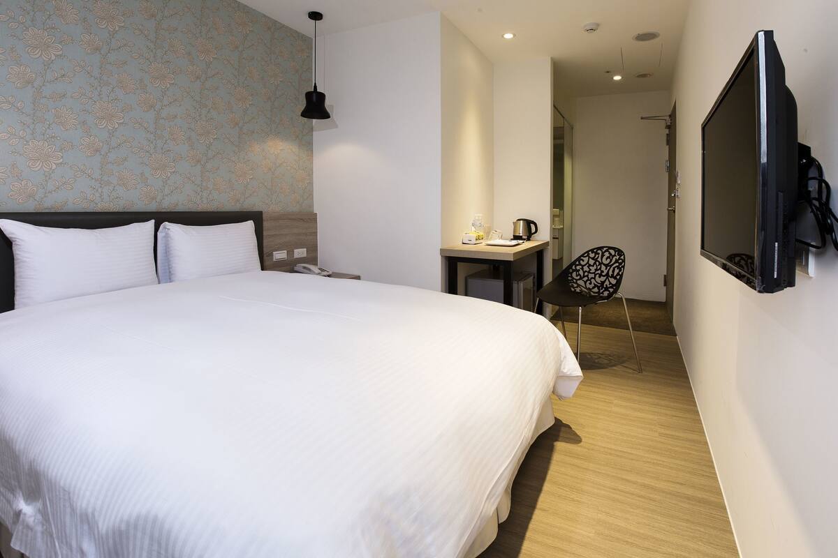 A hotel room with a bed, bedside table, chair, TV, and a small desk with a kettle.