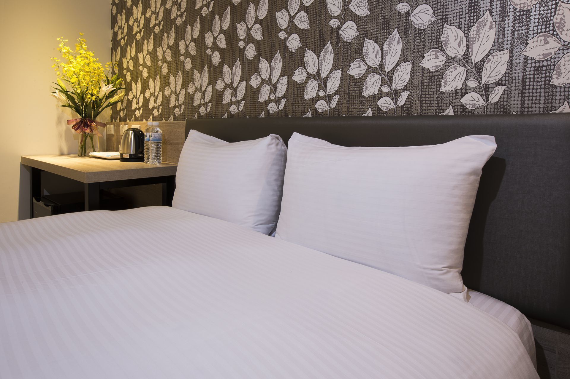 A hotel room with a bed, two pillows, a nightstand with a vase of flowers, a kettle, and water bottles.