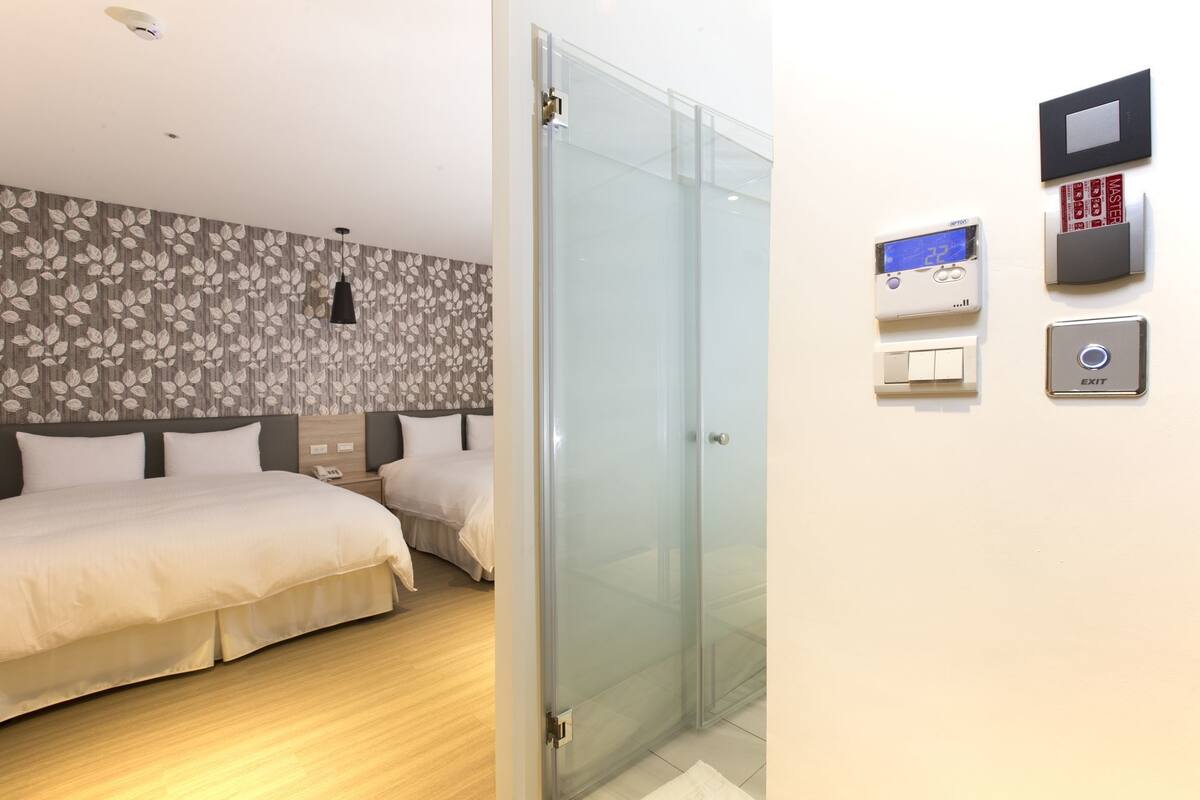 A hotel room with two beds, a glass door, and a thermostat on the wall.