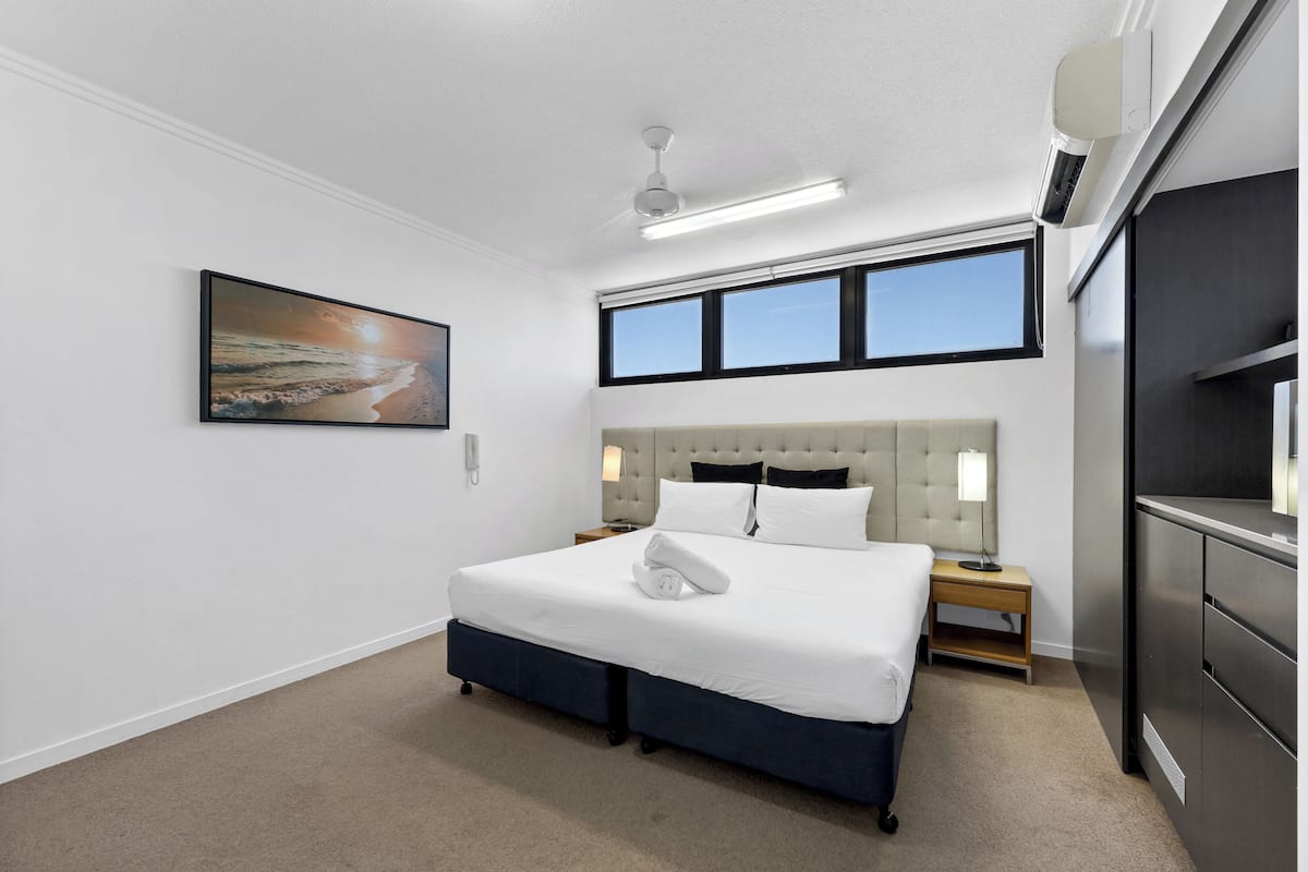A modern hotel room with a large bed, a headboard, a nightstand, a lamp, a ceiling fan, and a window with a view of the beach.