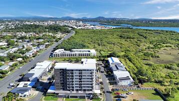 A modern multi-story building with a flat roof, surrounded by a parking area and lush greenery.