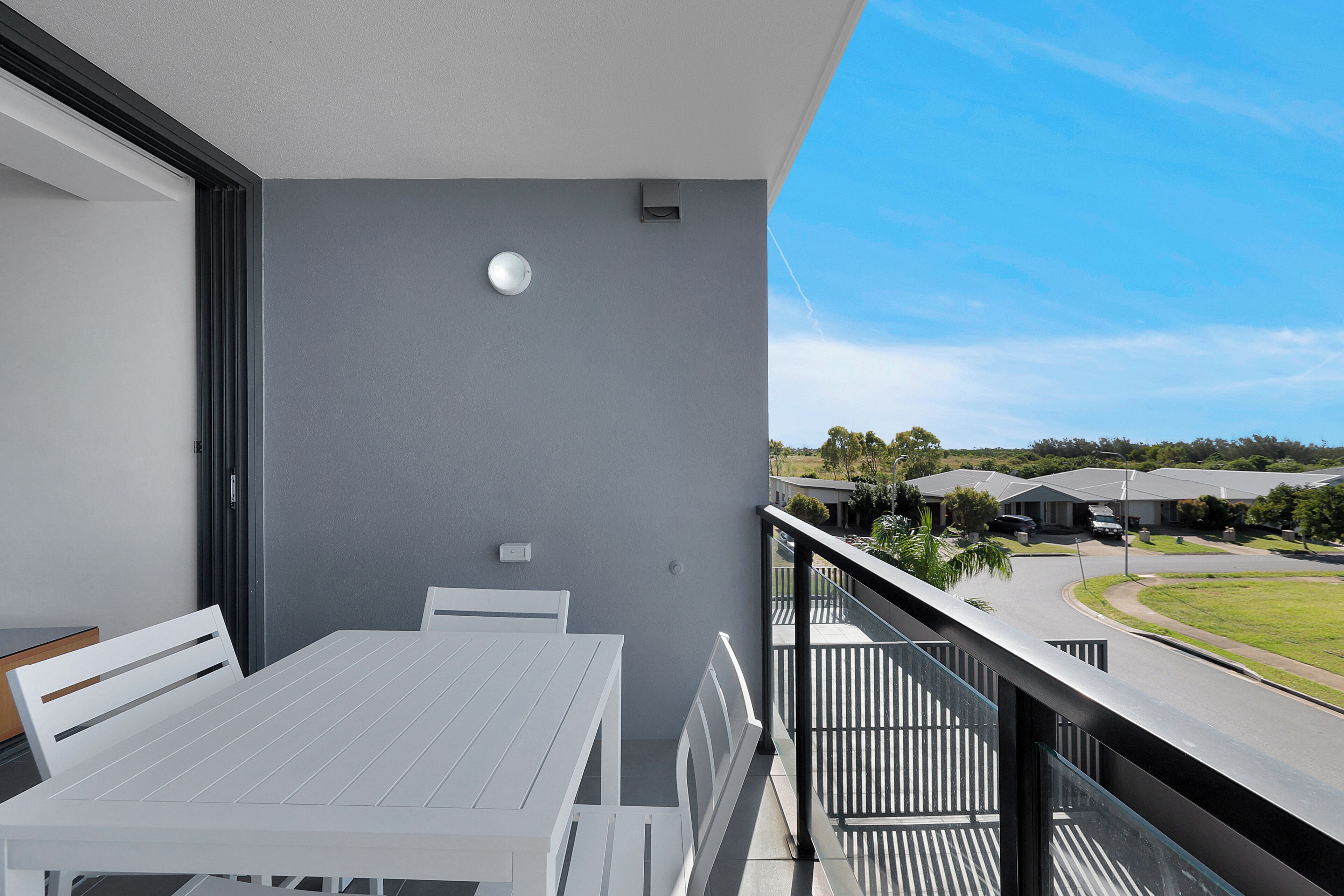 Een balkon met een witte tafel en stoelen, een grijze muur en uitzicht op een woongebied.