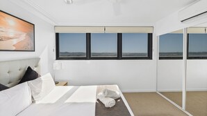 Et hotelværelse med en stor seng, udsigt til stranden gennem vinduet og en skydedør, der fører ud til en balkon.