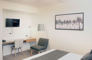 Una habitación de hotel moderna con cama, escritorio con silla, televisor, lámpara y un cuadro con palmeras.