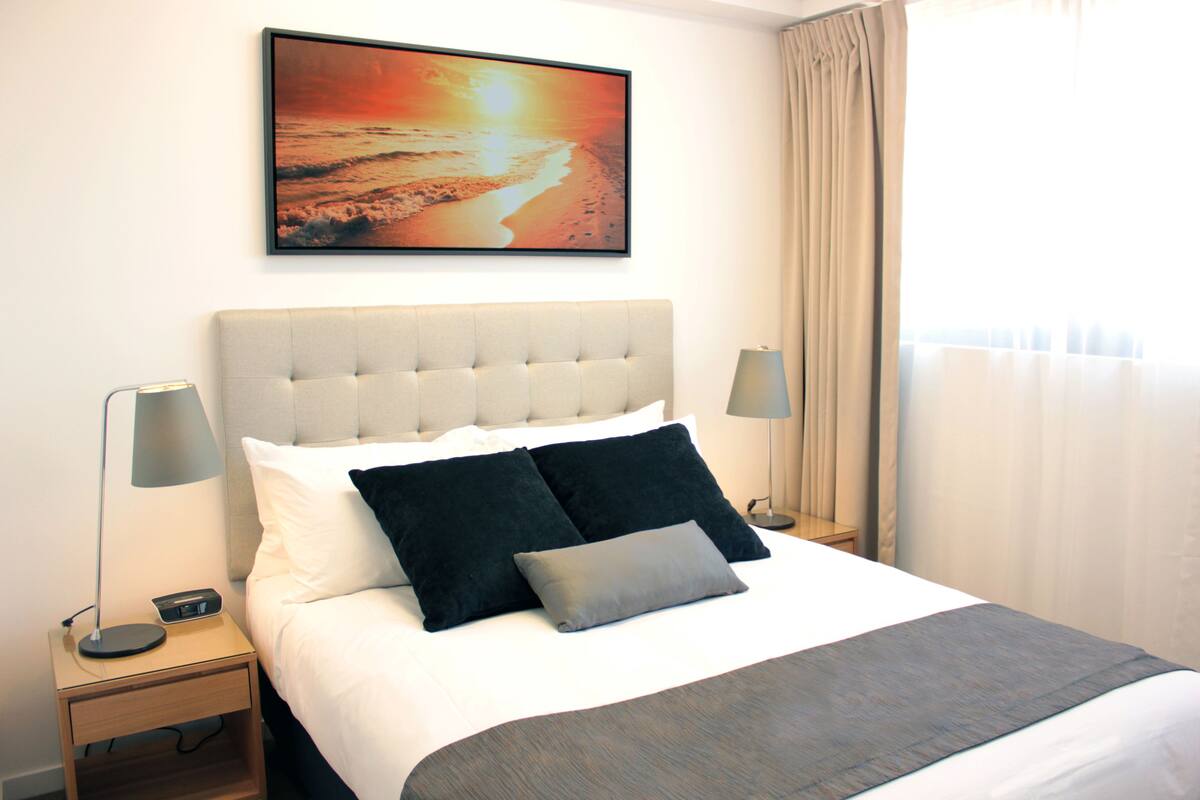 A neatly made bed with a grey and white duvet, two black pillows, and a grey throw. There is a framed picture of a sunset on the wall above the bed, and two bedside lamps on either side.