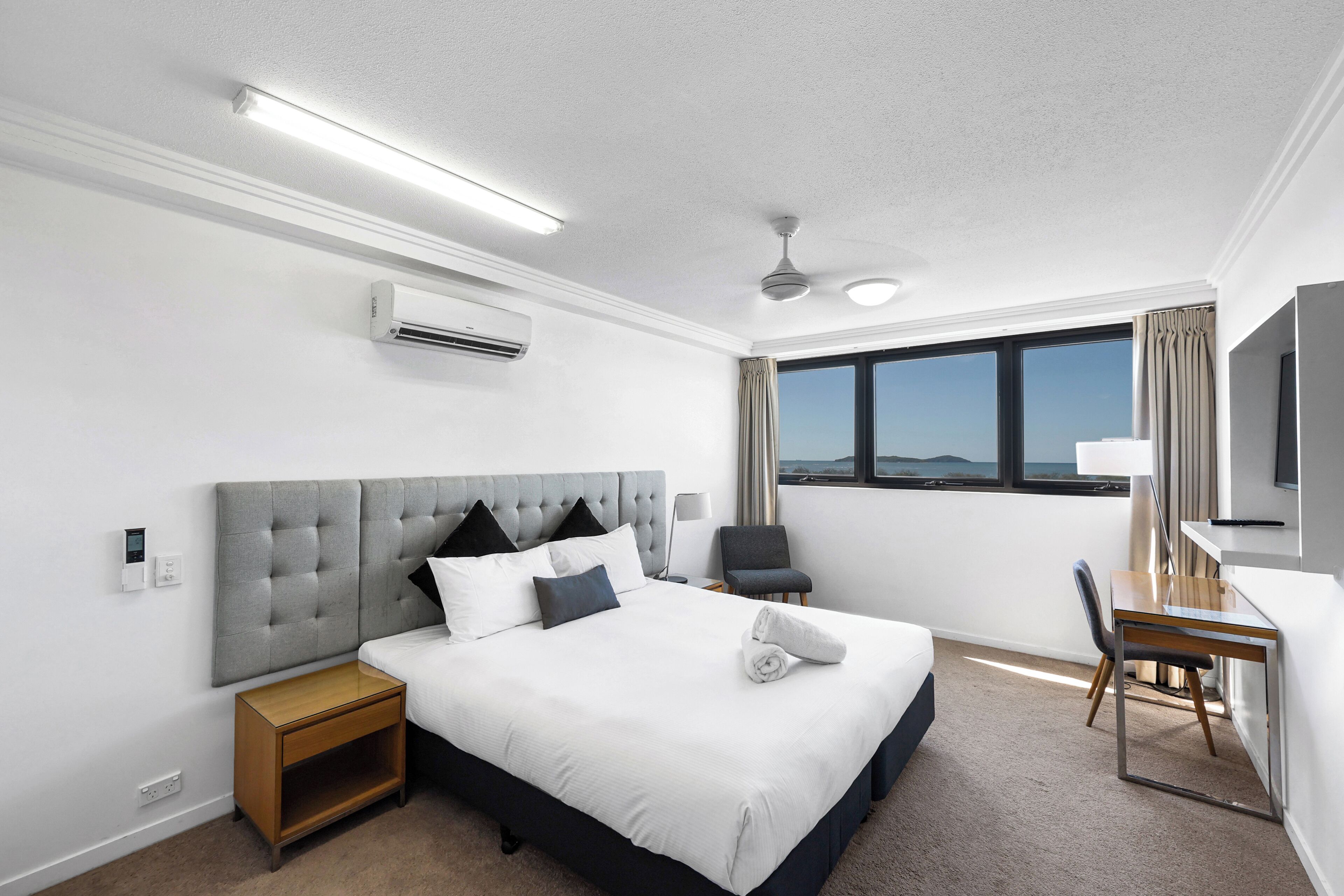 A hotel room with a large bed, a desk, a chair, a ceiling fan, and a large window offering a view.