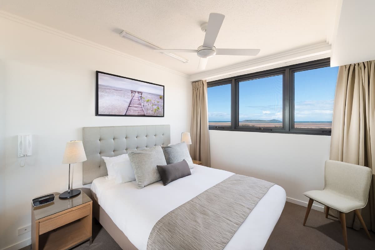 A hotel room with a bed, a chair, a nightstand, a ceiling fan, a window with a view, and a framed picture on the wall.