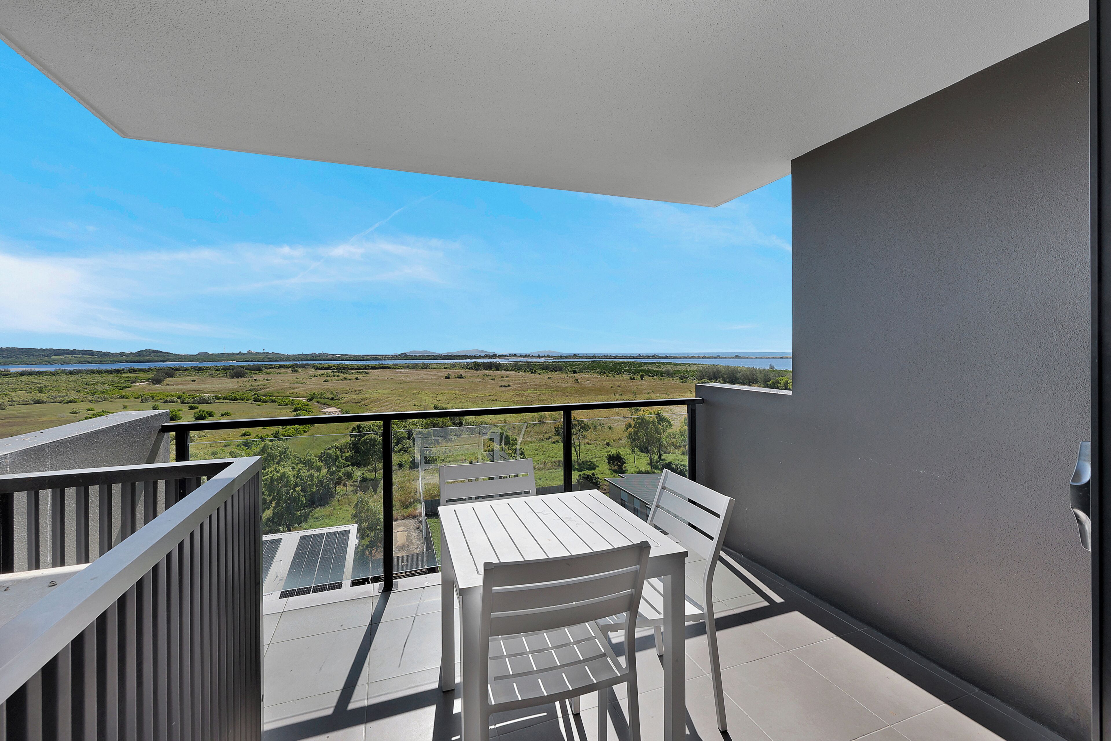 A balcony with a white table and chairs, offering a view of a green landscape and distant hills.