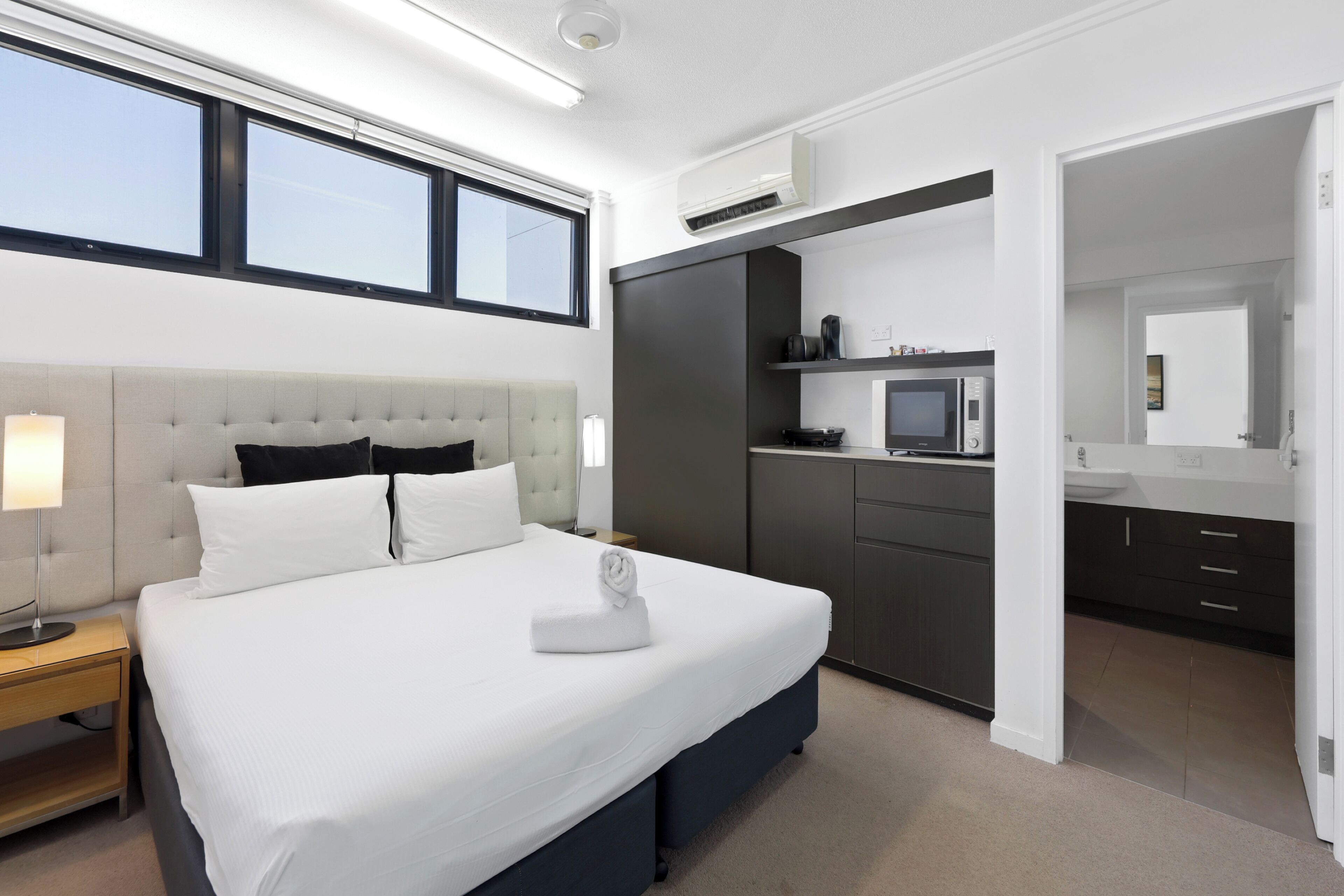 A modern hotel room with a large bed, a bedside table, a nightstand, a wardrobe, a kitchenette, and a bathroom visible through an open door.