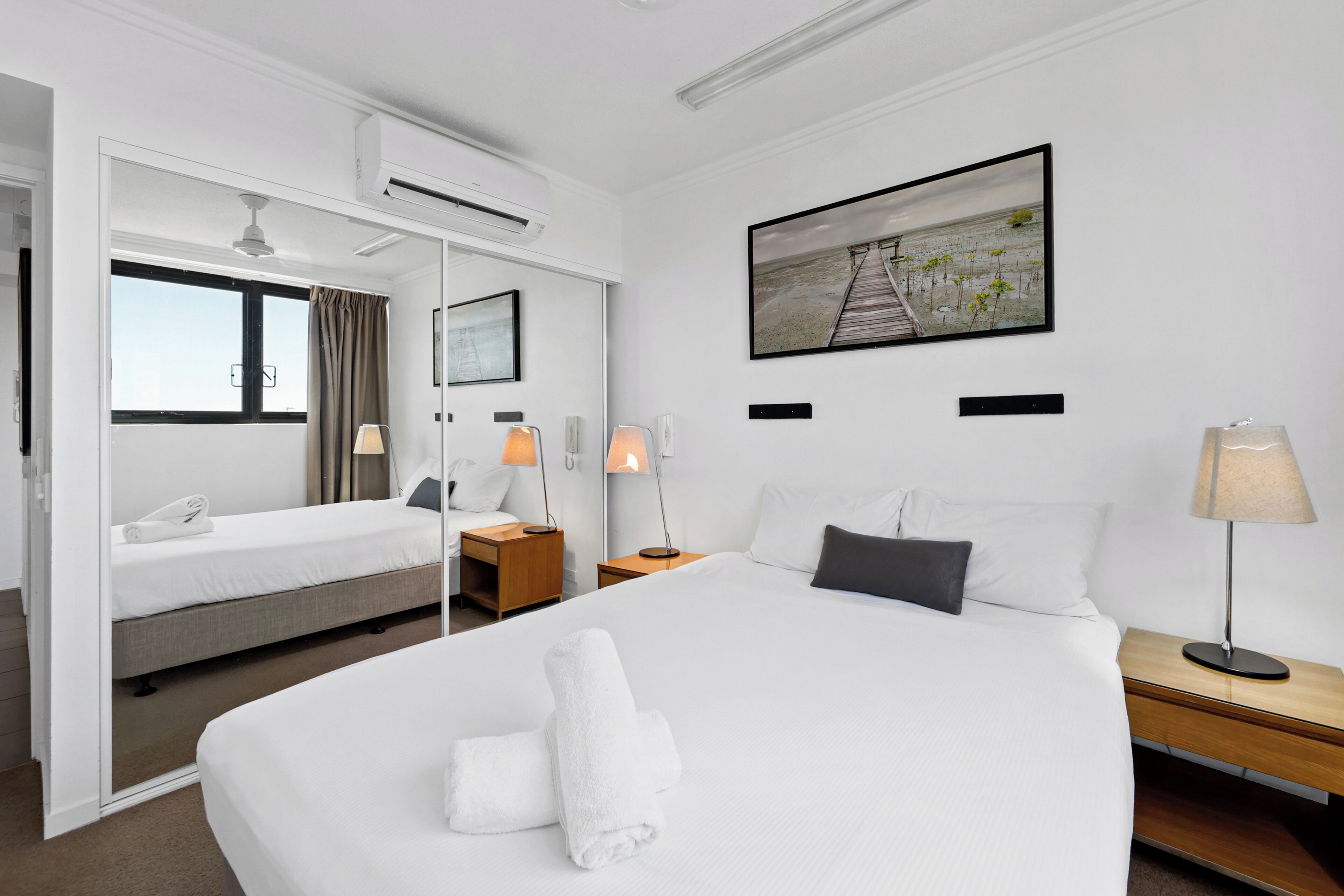 Una habitación de hotel con una cama grande, una mesita de noche, cortinas en la ventana, un equipo de aire acondicionado y un cuadro en la pared.