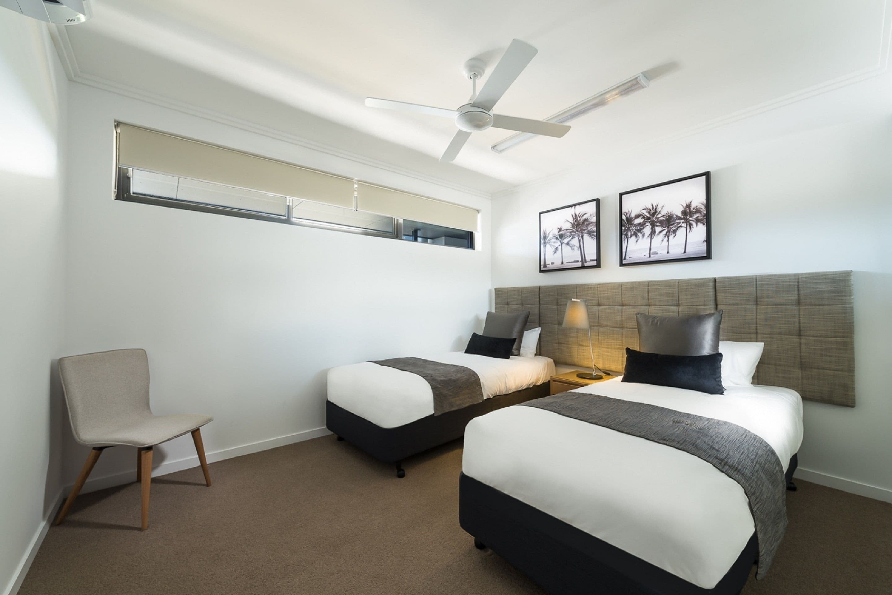 A hotel room with two beds, a ceiling fan, a window with blinds, and two framed pictures on the wall.