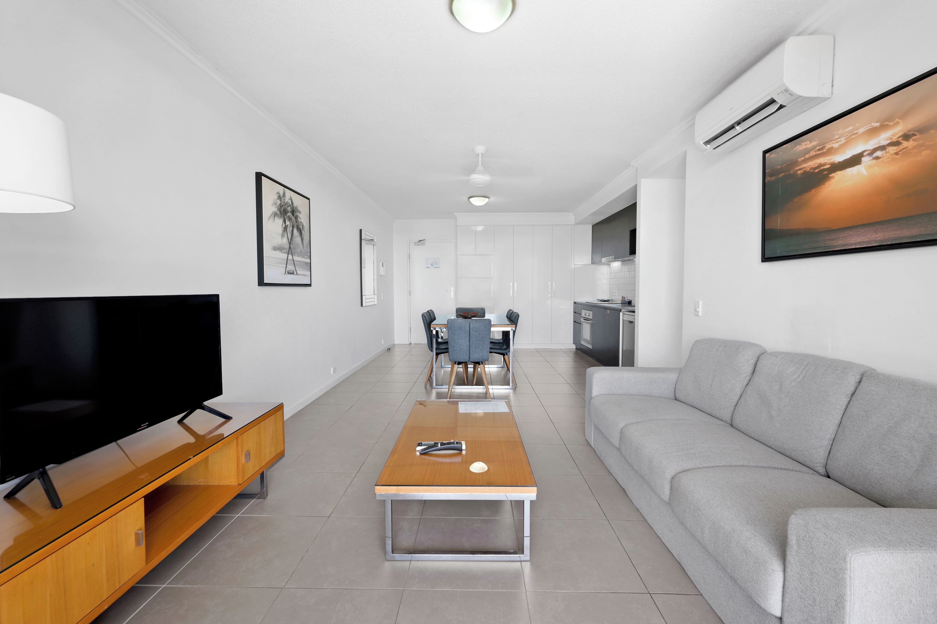 Ein modernes Wohnzimmer mit Flachbildfernseher, Couchtisch aus Holz, grauem Sofa und Essbereich mit Stühlen und Tisch.