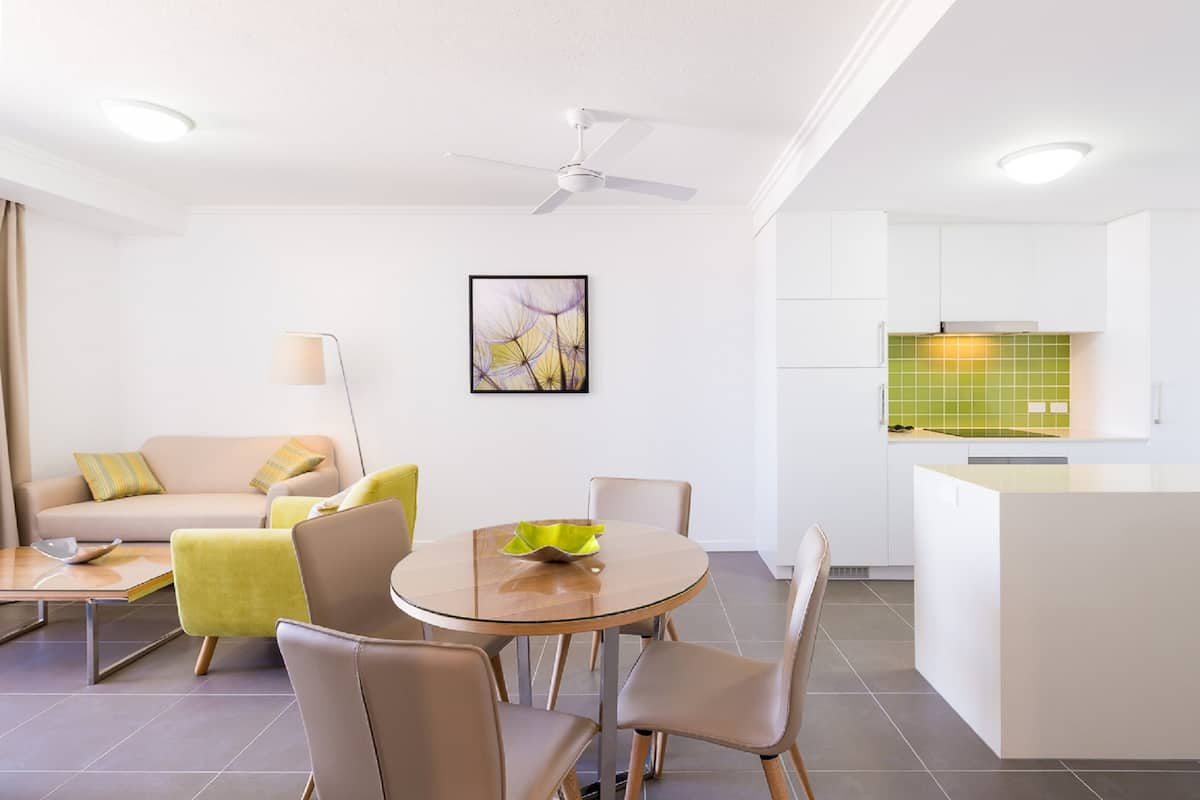 A modern living room with a round glass table, beige sofa, and yellow armchairs. A ceiling fan and recessed lighting are visible.