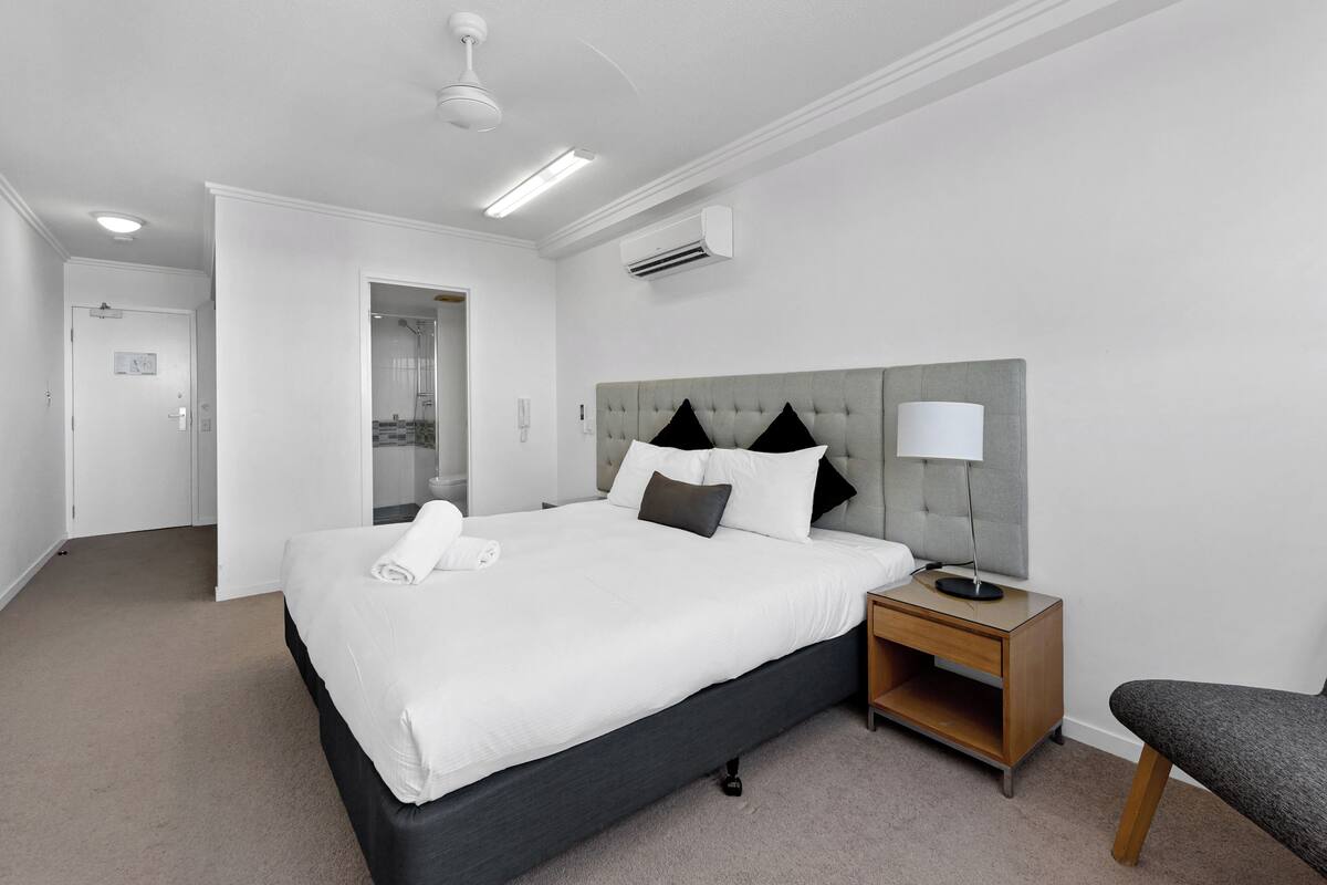 A hotel room with a large bed, a bedside table with a lamp, a chair, and a door leading to a bathroom.