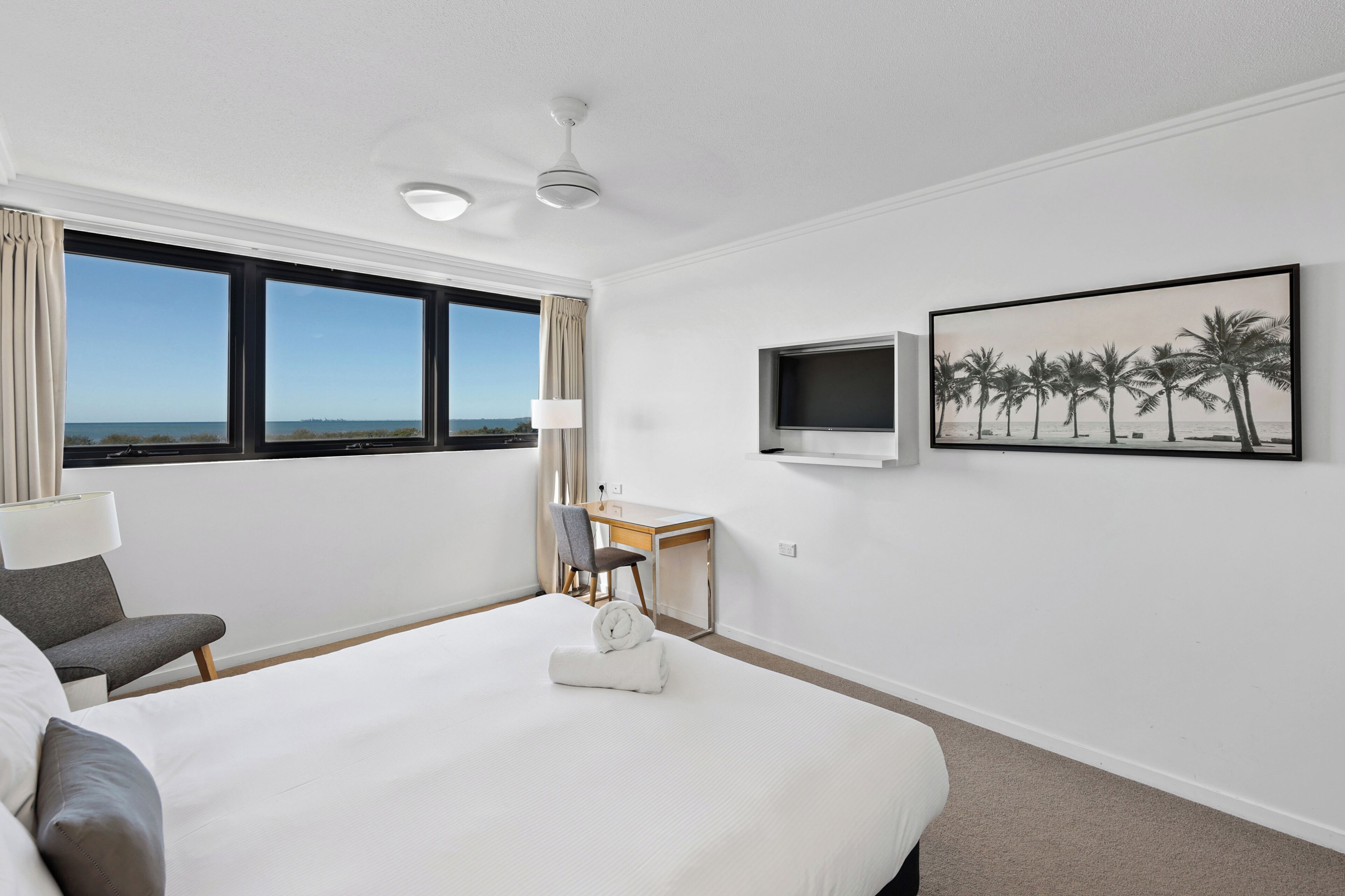 A hotel room with a large bed, a desk, a chair, a TV, and a window with a view of the sea.