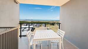 En balkon med et hvidt bord og stole, samt udsigt til havet og bygninger i det fjerne.