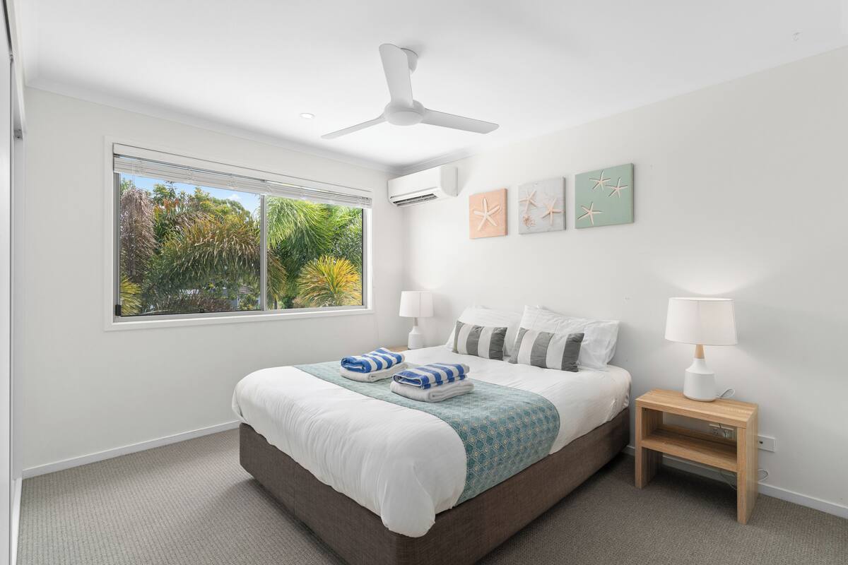 Une chambre à coucher avec un lit, un ventilateur de plafond, une table de chevet avec une lampe et une fenêtre donnant sur un paysage verdoyant.