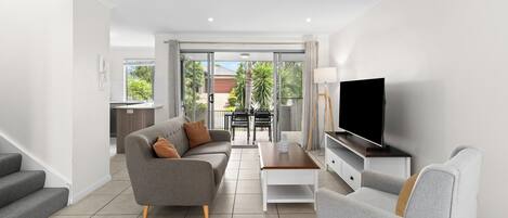 A modern living room with a grey sofa, a coffee table, a flat-screen TV, and a staircase leading to an outdoor area.