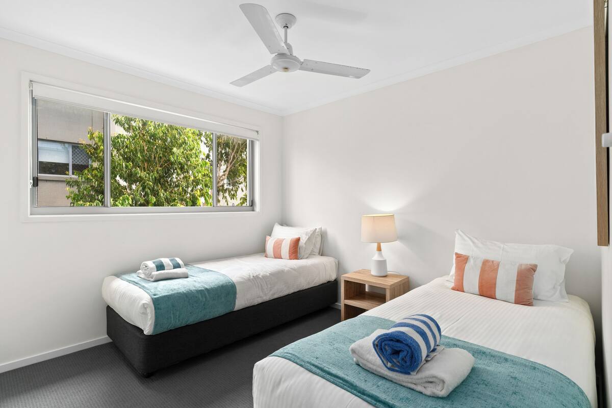 Une chambre d’hôtel avec deux lits, un ventilateur de plafond, une fenêtre donnant sur la verdure et une table de chevet avec une lampe.