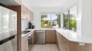 Una cocina moderna con lavavajillas, horno y refrigerador de acero inoxidable. Cuenta con gabinetes de madera, encimera blanca y un ventanal con vistas al exterior.