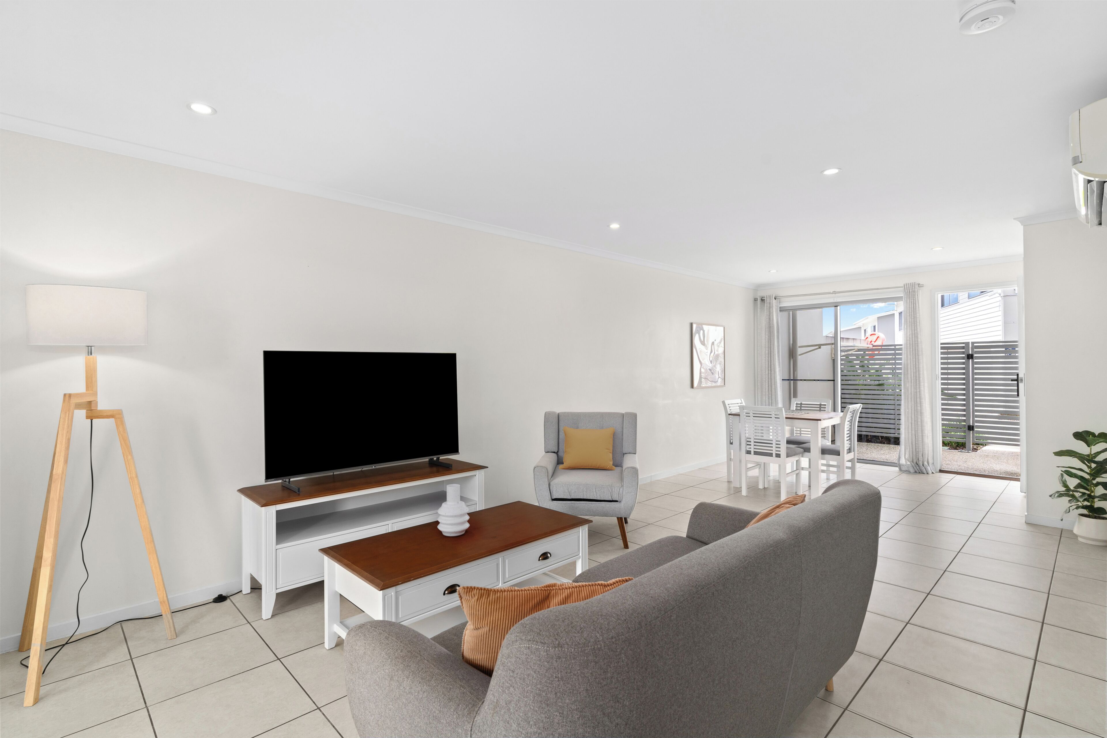 Un salon moderne doté d’un téléviseur à écran plat, d’une table basse en bois, d’un canapé gris, d’un fauteuil blanc et d’un coin repas avec une table et des chaises.