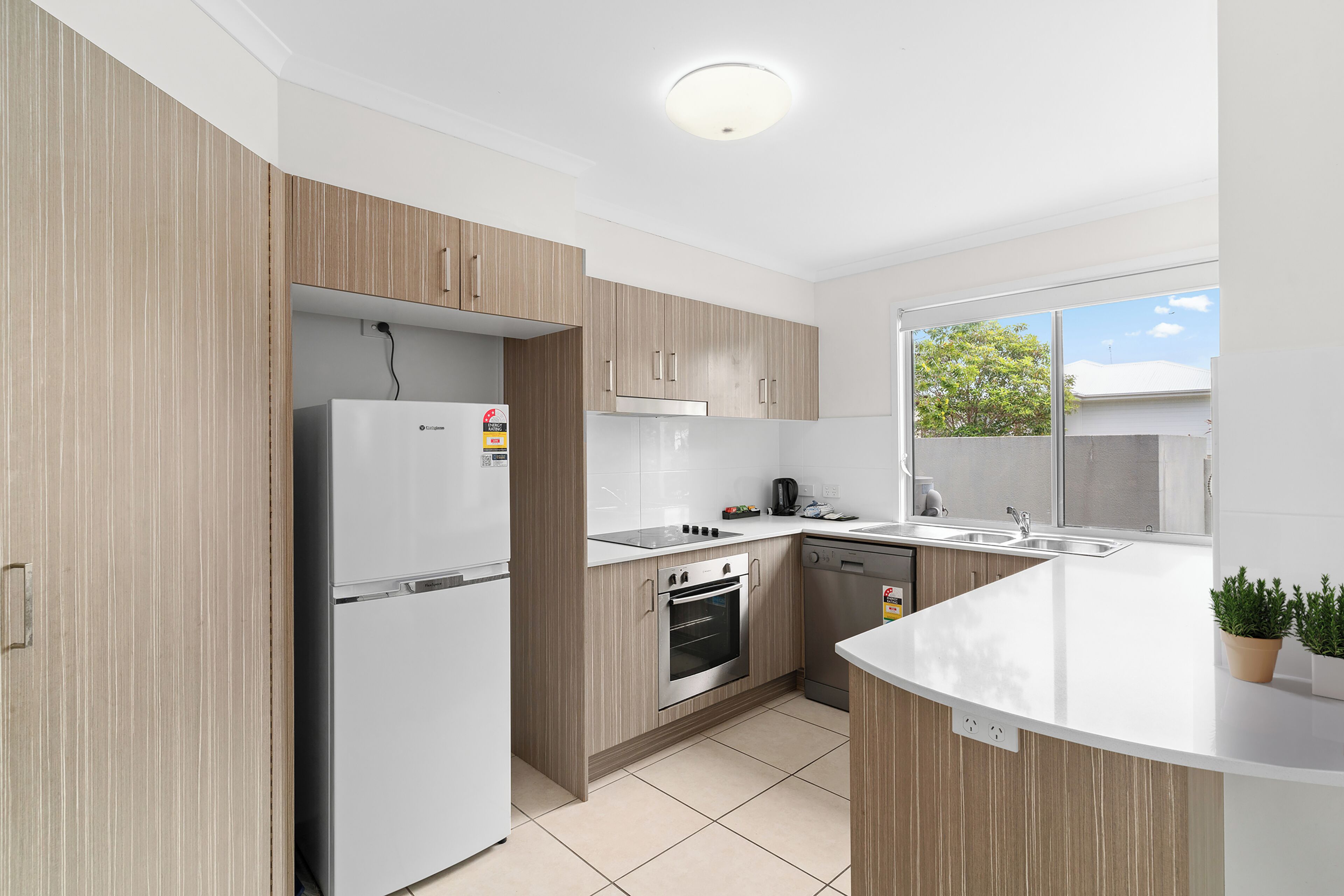 A modern kitchen with a refrigerator, oven, dishwasher, and a window with a view of trees.