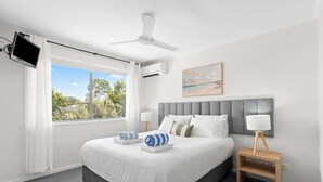 Un dormitorio moderno con una cama grande, un ventilador de techo, un televisor montado en la pared y una ventana con vistas a árboles.