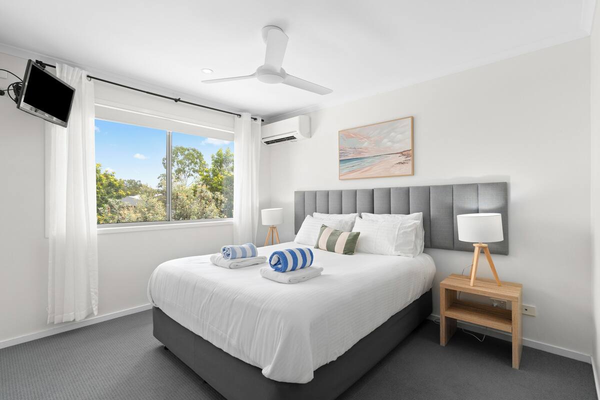 Une chambre moderne avec un grand lit, un ventilateur de plafond, un téléviseur fixé au mur et une fenêtre donnant sur la verdure.