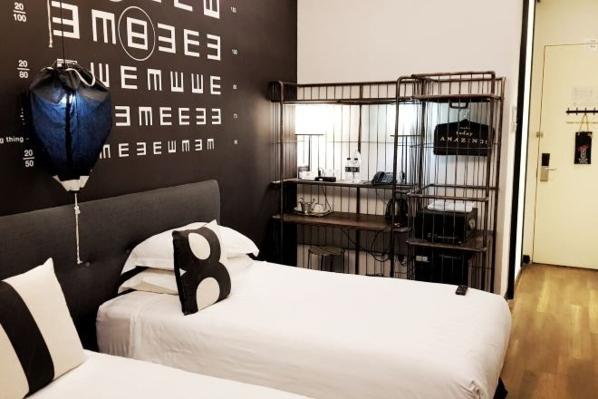 A hotel room with a bed, a wall-mounted shelf with a hanging shirt, and a window with blinds.