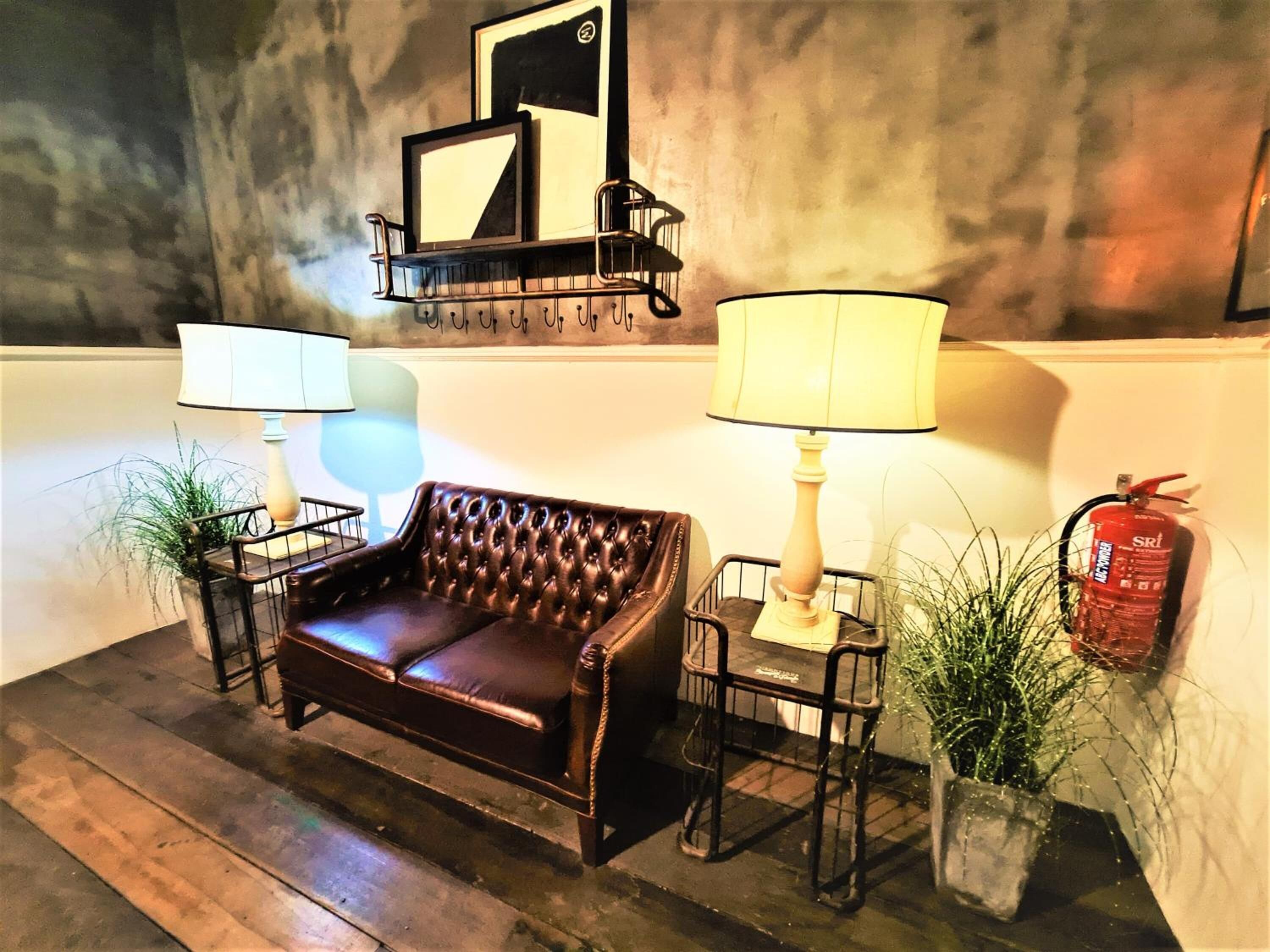 A dark brown leather sofa in a room with two floor lamps, a fire extinguisher, and a wall-mounted shelf with framed pictures.