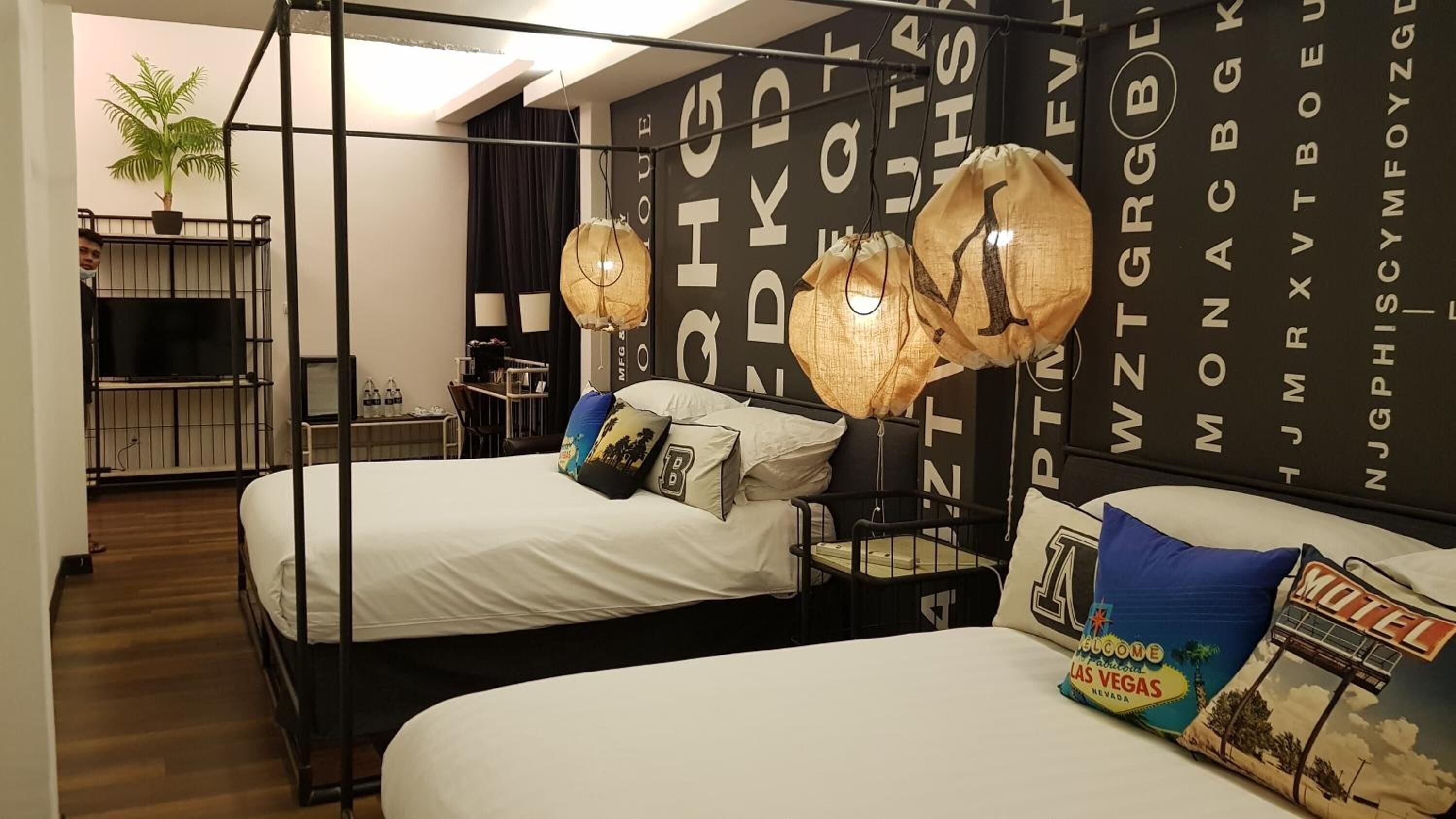 Habitación de hotel con dos camas, una planta grande en maceta, un escritorio y una pared negra con texto blanco.