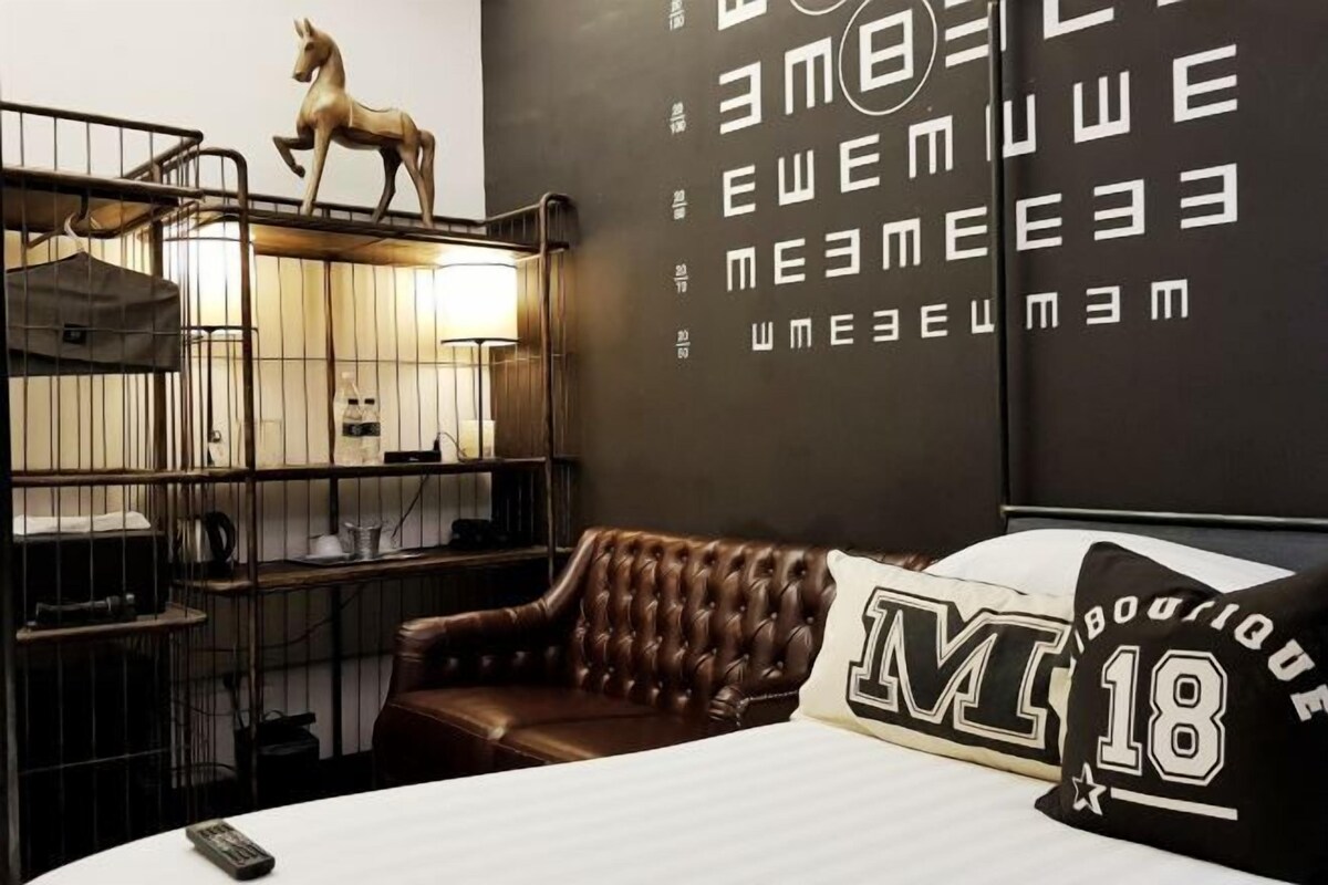 A hotel room with a bed, a brown leather armchair, a small wooden horse sculpture on a shelf, and a wall with repeating text patterns.